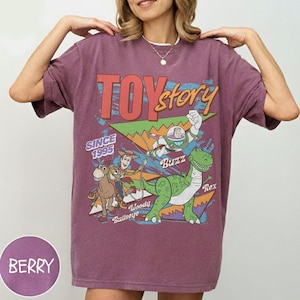 May include: Berry-coloured t-shirt with a "Toy Story" graphic. The design includes the text "TOY STORY" and the year "SINCE 1995." Characters Woody, Buzz, Rex, and Bullseye are also featured in the graphic, in a vintage style.