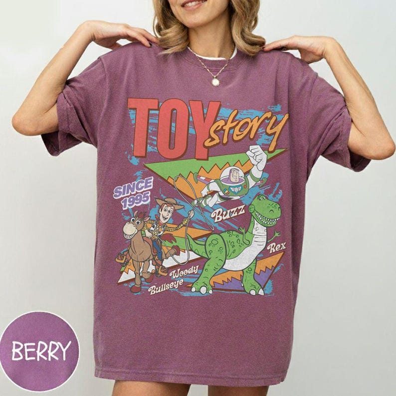 May include: Berry-coloured t-shirt with a "Toy Story" graphic. The design includes the text "TOY STORY" and the year "SINCE 1995." Characters Woody, Buzz, Rex, and Bullseye are also featured in the graphic, in a vintage style.