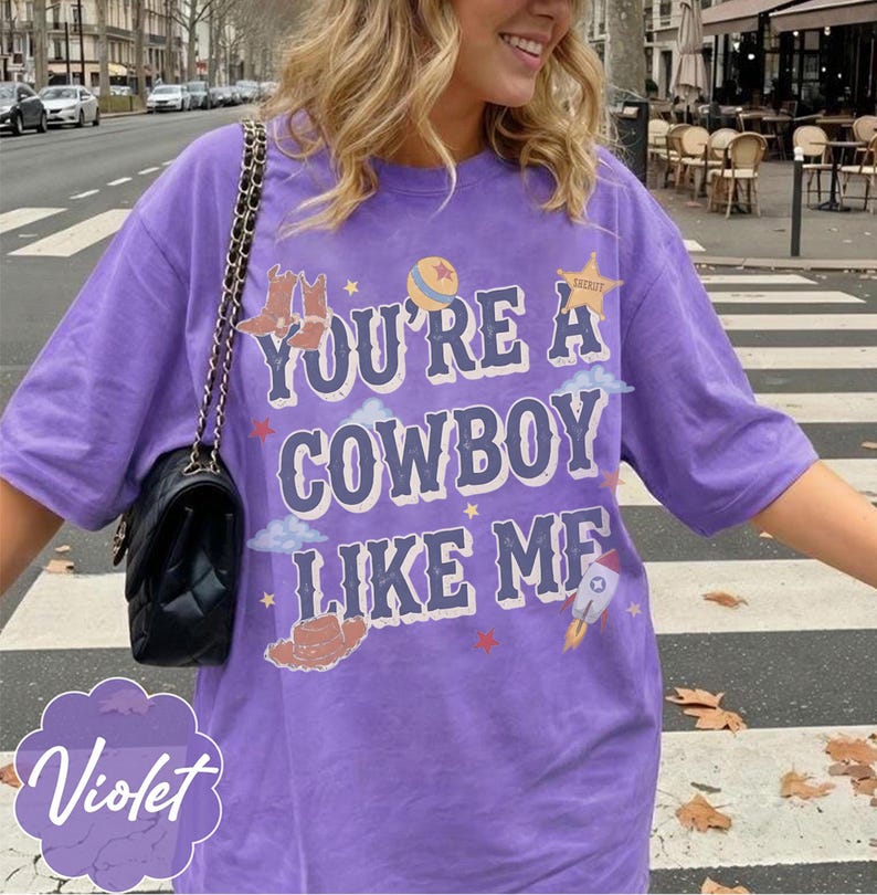 May include: A purple short-sleeve t-shirt featuring the text "YOU'RE A COWBOY LIKE ME" in a distressed font. The graphic includes illustrations of cowboy boots, a ball, a sheriff star, clouds, a cowboy hat, and a rocket ship with small stars.