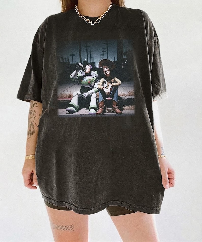 Puede incluir: Camiseta gris oscuro con un gr&aacute;fico de Buzz Lightyear y Woody de Toy Story. La imagen muestra a los personajes sentados juntos en una escena callejera. La camiseta tiene un aspecto vintage y desgastado.