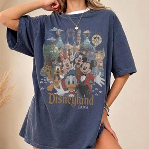 May include: A navy blue short-sleeve t-shirt featuring a graphic print of Mickey Mouse, Minnie Mouse, Donald Duck, Goofy, and Pluto in front of a castle. The text reads Disneyland Est. 1955. The shirt has a crew neckline and a relaxed, loose fit.