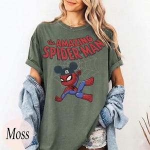 May include: Moss green t-shirt featuring a cartoon Spider-Man character with Mickey Mouse ears, swinging from a web. The text "the AMAZING SPIDER-MAN" is arched above the character in red. The shirt is a relaxed fit.