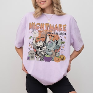 Puede incluir: Camiseta lila claro con un gr&aacute;fico de Halloween. El dise&ntilde;o incluye el texto "Nightmare on Main Street" y personajes de dibujos animados. Tambi&eacute;n se ven un fantasma, calabazas y una l&aacute;pida.