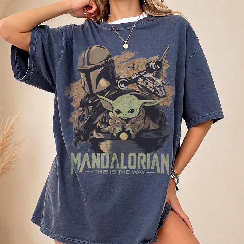 May include: Navy blue t-shirt featuring a graphic of the Mandalorian, Grogu, and a spaceship. The text "MANDALORIAN - THIS IS THE WAY" is printed below the image. The shirt has short sleeves and a relaxed fit.