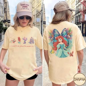 May include: A light yellow t-shirt featuring a front graphic with cartoon characters and text that reads "I want to be where the people are." The back of the shirt has a graphic of a mermaid. The shirt is a comfortable, casual style.