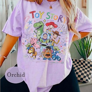 Puede incluir: Camiseta color orqu&iacute;dea con un gr&aacute;fico colorido de personajes de Toy Story. El dise&ntilde;o incluye las palabras "Toy Story" en rojo y naranja, junto con ilustraciones de Woody, Buzz Lightyear y otros personajes. La camiseta tiene mangas cortas.