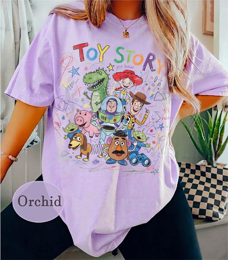 Puede incluir: Camiseta color orqu&iacute;dea con un gr&aacute;fico colorido de personajes de Toy Story. El dise&ntilde;o incluye las palabras "Toy Story" en rojo y naranja, junto con ilustraciones de Woody, Buzz Lightyear y otros personajes. La camiseta tiene mangas cortas.