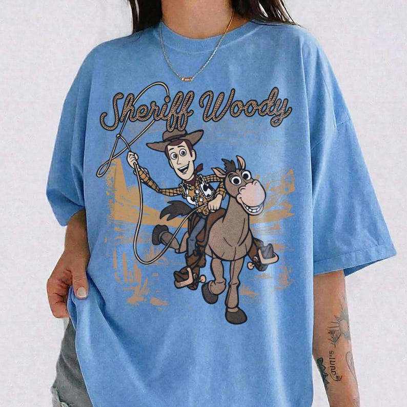 May include: A light blue t-shirt featuring a graphic print of the character Sheriff Woody riding a horse while holding a lasso. The text "Sheriff Woody" is printed in a brown, rope-style font above the character illustration.