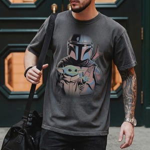 May include: A charcoal gray short-sleeve t-shirt featuring a graphic print of a helmeted warrior in silver armor standing behind a small green creature with large ears and a tan robe. The shirt has a crew neckline and a relaxed fit.