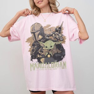 May include: A light pink t-shirt featuring a graphic of The Mandalorian and Grogu from the Star Wars series. The design includes the text "MANDALORIAN - THIS IS THE WAY" in a green font. The shirt is a casual, oversized style.