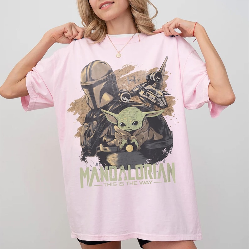 May include: A light pink t-shirt featuring a graphic of The Mandalorian and Grogu from the Star Wars series. The design includes the text "MANDALORIAN - THIS IS THE WAY" in a green font. The shirt is a casual, oversized style.