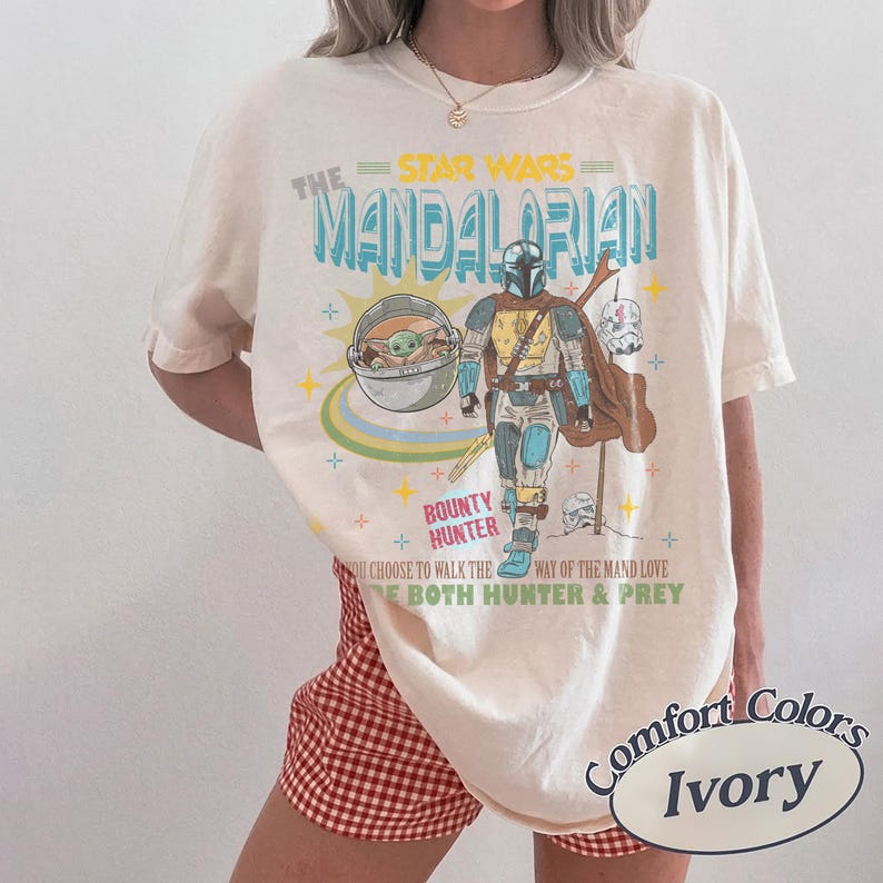 May include: Ivory-colored t-shirt featuring a graphic of The Mandalorian, Grogu, and text that reads "Bounty Hunter." The design includes the Star Wars logo and the phrase "You choose to walk the way of the Mand love, be both hunter & prey."