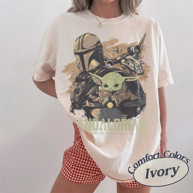 May include: Ivory-colored t-shirt featuring a graphic of The Mandalorian, Grogu, and a spaceship. The text "MANDALORIAN" and "THIS IS THE WAY" are printed below the image. The shirt is a Comfort Colors brand.