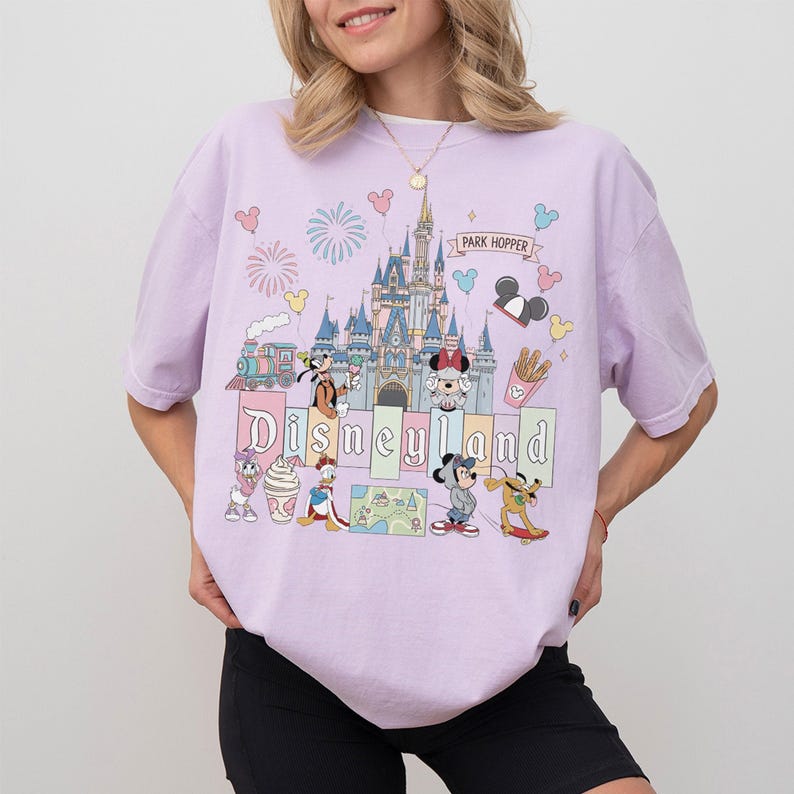 May include: A light purple t-shirt featuring a Disneyland graphic. The graphic includes the Disneyland castle, various Disney characters, fireworks, and the words "Disneyland" and "Park Hopper".