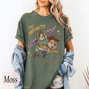 May include: A moss green crew neck t-shirt featuring a graphic of two chipmunks dressed as space and cowboy characters. The text reads TO INFINITY AND BEYOND. The shirt has short sleeves and a relaxed fit, shown with a denim jacket draped over the arms.