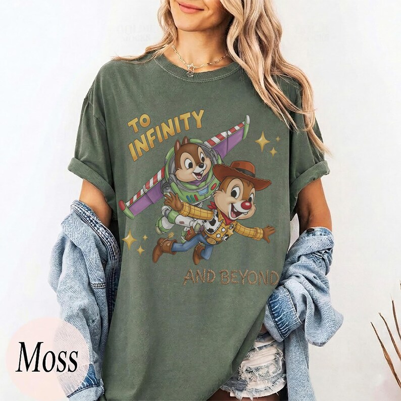 May include: A moss green crew neck t-shirt featuring a graphic of two chipmunks dressed as space and cowboy characters. The text reads TO INFINITY AND BEYOND. The shirt has short sleeves and a relaxed fit, shown with a denim jacket draped over the arms.