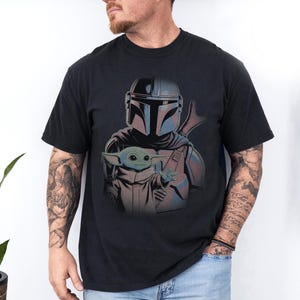 May include: A black short-sleeved t-shirt featuring a graphic print of a Mandalorian character in silver armor standing behind a small green creature with large ears and a tan robe. The design is centered on the chest of the garment.