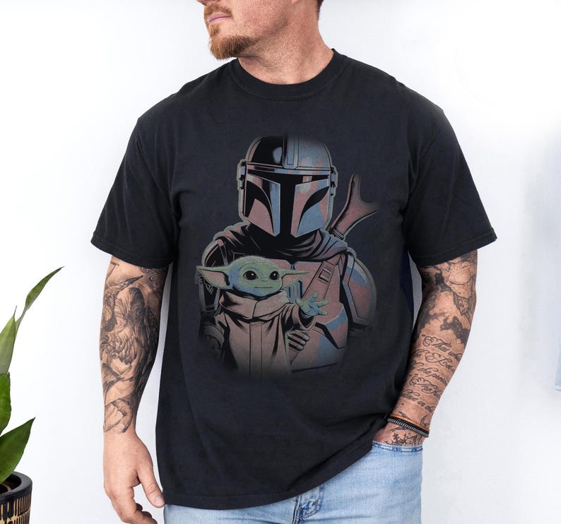 May include: A black short-sleeved t-shirt featuring a graphic print of a Mandalorian character in silver armor standing behind a small green creature with large ears and a tan robe. The design is centered on the chest of the garment.