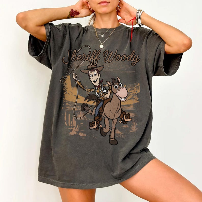 May include: A charcoal gray short-sleeve t-shirt featuring a graphic print of Sheriff Woody riding a horse. The text Sheriff Woody is printed in a brown cursive font above the character. The design includes a lasso and a desert landscape background.