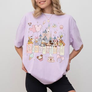 May include: A lilac t-shirt with a "Disneyland" graphic. The design includes cartoon characters such as Mickey and Minnie Mouse, Donald Duck, and Pluto, along with a castle, balloons, and desserts. The shirt is a pastel colour.