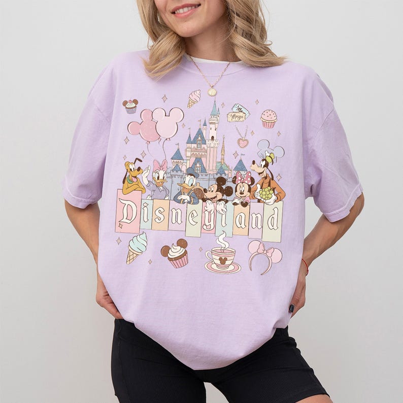 May include: A lilac t-shirt with a "Disneyland" graphic. The design includes cartoon characters such as Mickey and Minnie Mouse, Donald Duck, and Pluto, along with a castle, balloons, and desserts. The shirt is a pastel colour.