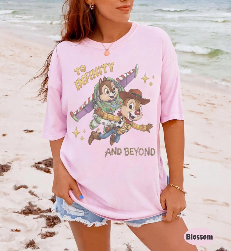 May include: A light pink short-sleeved t-shirt featuring a graphic of two chipmunks dressed as space and cowboy characters. The text reads TO INFINITY AND BEYOND. The design includes small yellow star accents and the characters are depicted in flight.