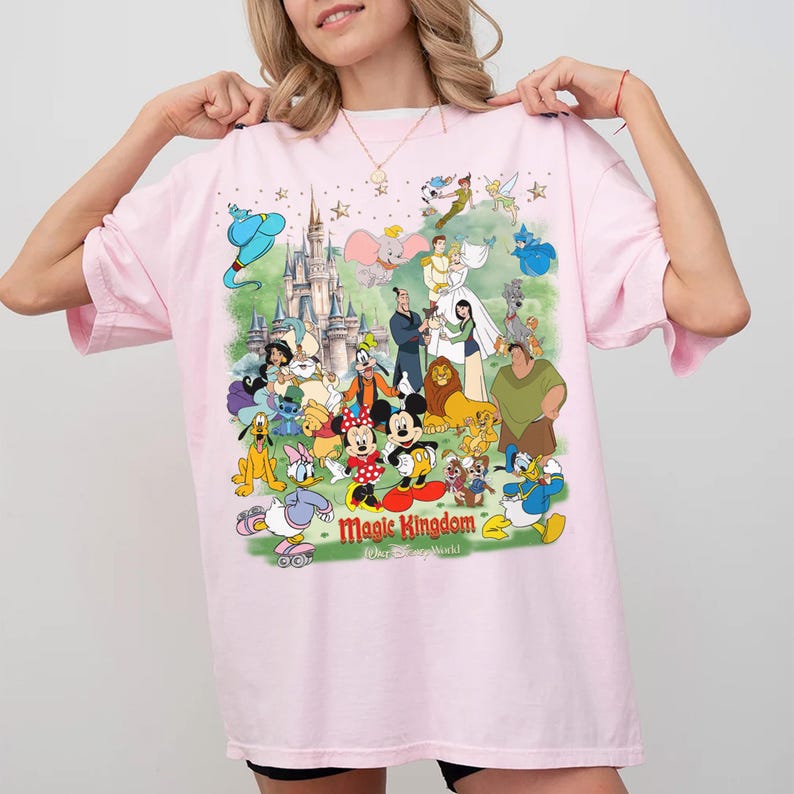May include: Pale pink t-shirt with a vibrant graphic of Disney characters such as Mickey Mouse, Donald Duck, and Cinderella. The words "Magic Kingdom" are printed below the characters. The shirt has short sleeves and a round neck.