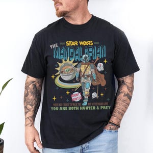 May include: Black t-shirt featuring a graphic from The Mandalorian. The design includes the text "The Star Wars Mandalorian", a depiction of the Mandalorian and Grogu, and the words "Bounty Hunter".