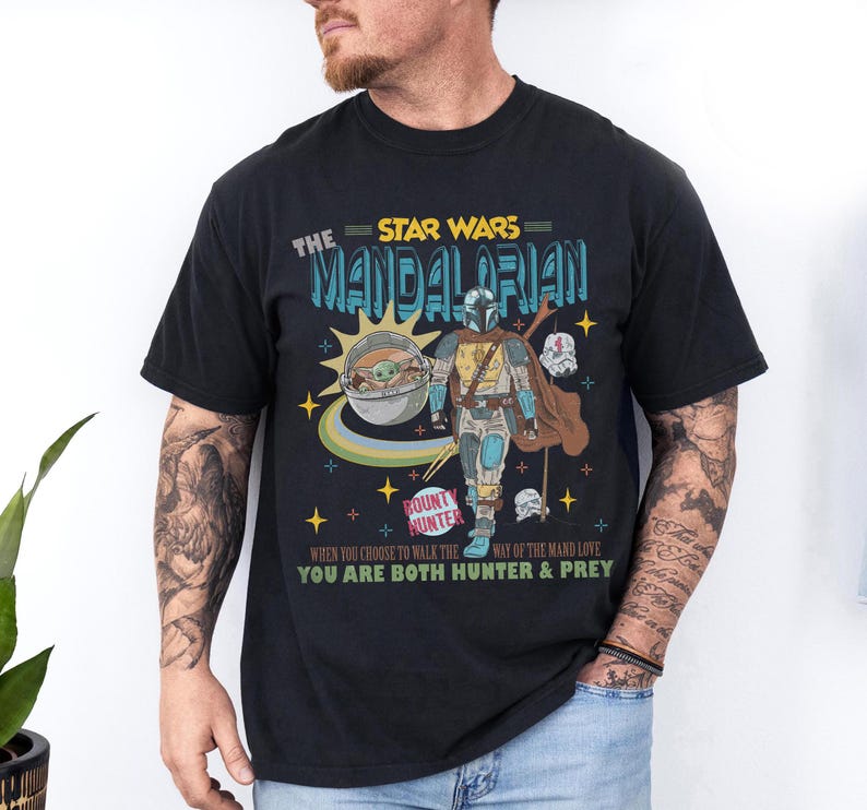 May include: Black t-shirt featuring a graphic from The Mandalorian. The design includes the text "The Star Wars Mandalorian", a depiction of the Mandalorian and Grogu, and the words "Bounty Hunter".