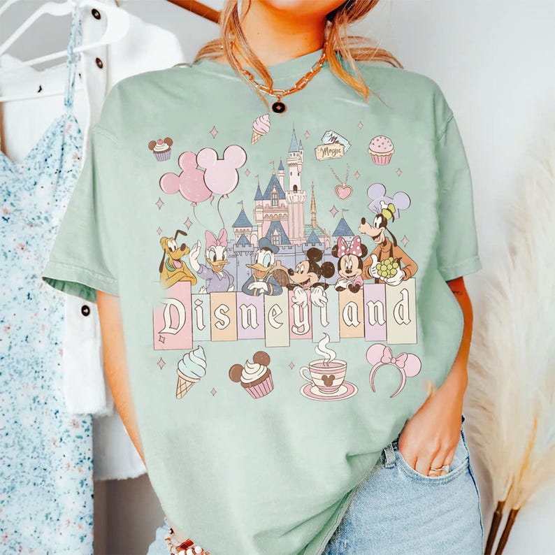 May include: A pale green t-shirt with the word "Disneyland" in pastel letters, featuring cartoon characters and a castle graphic. The shirt has a crew neck and short sleeves. Additional graphics include ice cream cones, cupcakes, and Mickey Mouse ears.