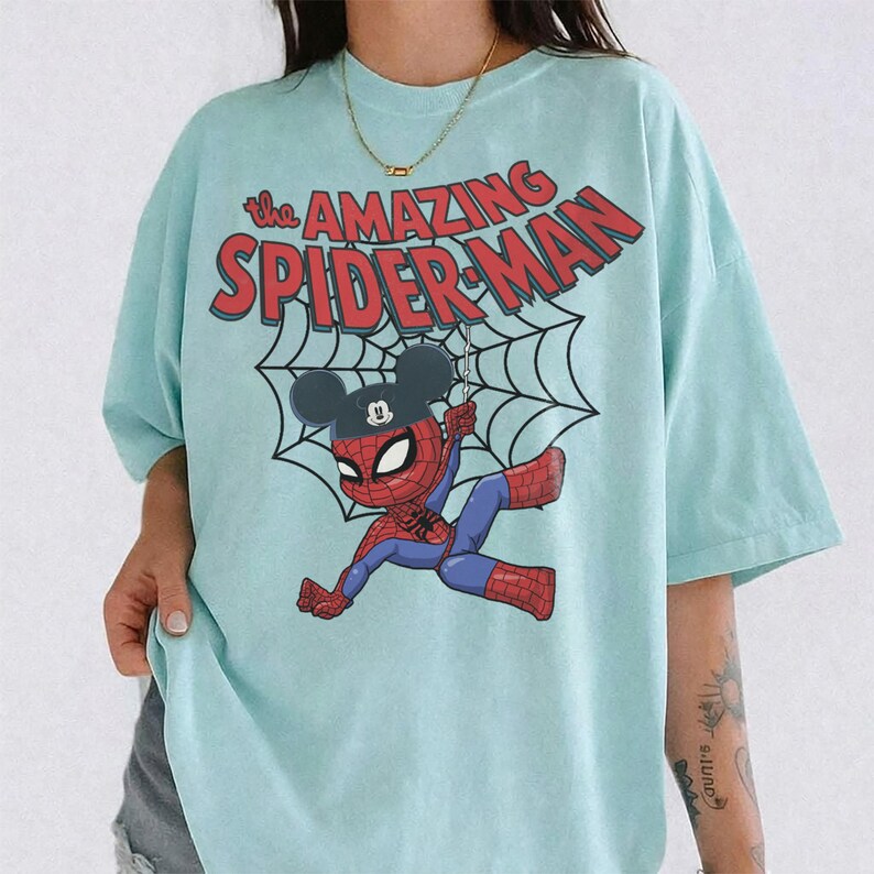 May include: Light blue t-shirt featuring a cartoon Spider-Man character with a Mickey Mouse hat hanging from a web. The text "The Amazing Spider-Man" is printed in red above the character.