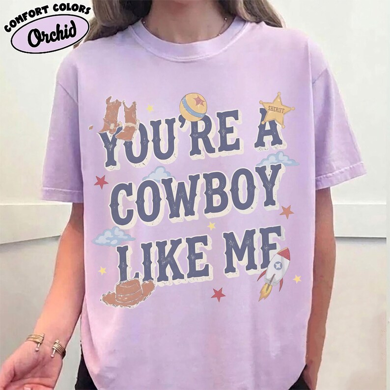 May include: A light purple short-sleeve t-shirt featuring the dark blue text "YOU'RE A COWBOY LIKE ME" surrounded by graphic illustrations of cowboy boots, a ball, a sheriff star, clouds, a hat, a rocket ship, and small stars.