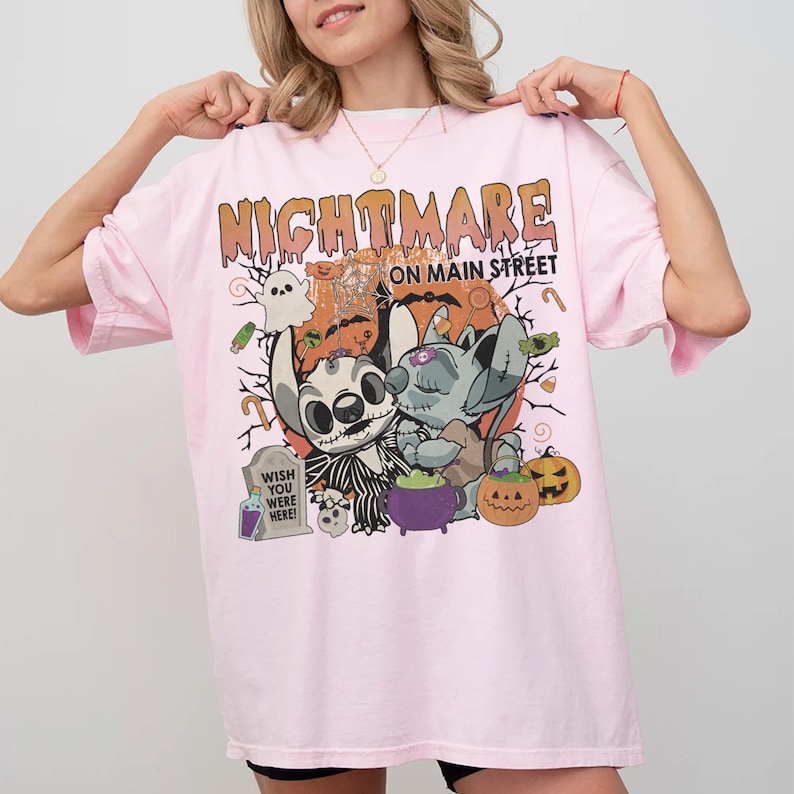 Puede incluir: Camiseta rosa claro con un dise&ntilde;o tem&aacute;tico de Halloween. El dise&ntilde;o incluye las palabras "NIGHTMARE ON MAIN STREET" en naranja, junto con personajes de dibujos animados, calabazas y una l&aacute;pida que dice "WISH YOU WERE HERE!"