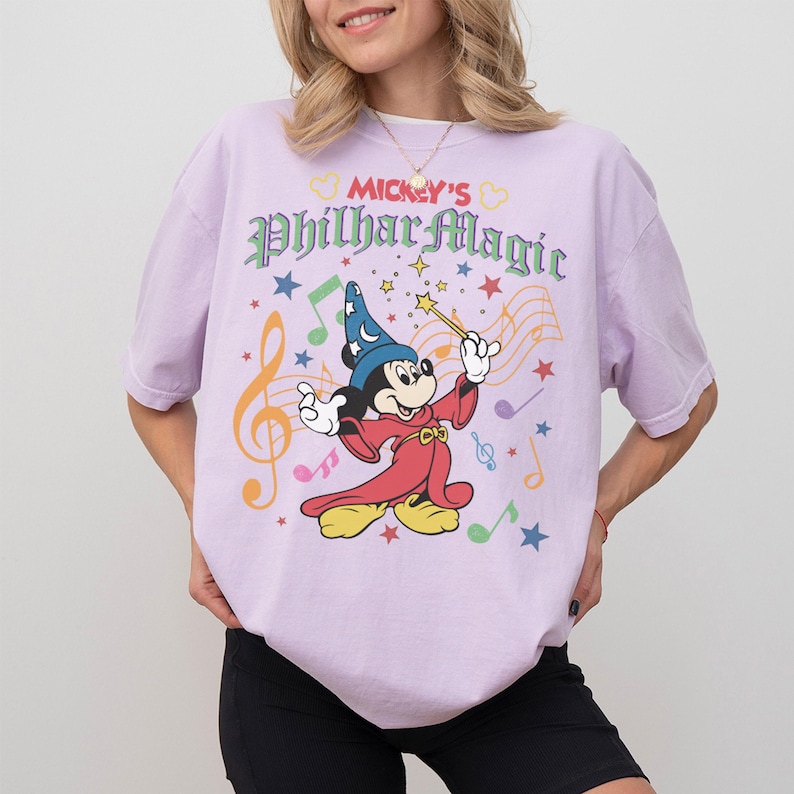 May include: A light purple t-shirt featuring Mickey Mouse in a sorcerer's outfit, holding a wand. The shirt has the text "Mickey's PhilharMagic" with musical notes and stars. The shirt is a casual wear item.