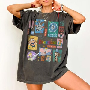 K&ouml;nnte beinhalten: Dunkelgraues T-Shirt mit einem farbenfrohen Disneyland-Motiv. Das Design zeigt verschiedene Zeichentrickfiguren und den Text "Disneyland" und "1955". Das T-Shirt hat einen Vintage-Look.