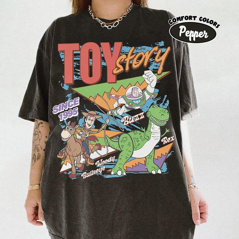 May include: Black t-shirt with a vibrant Toy Story graphic featuring Woody, Buzz Lightyear, and Rex. The design includes the text "TOY STORY" and "SINCE 1995". The shirt is labeled "Comfort Colors Pepper".