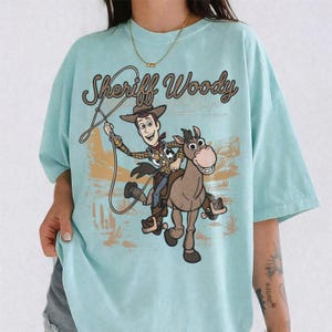 May include: A light blue t-shirt featuring a graphic print of Sheriff Woody riding a horse. The design includes the text Sheriff Woody in a brown rope-style font, with a desert landscape background in shades of brown and tan behind the characters.