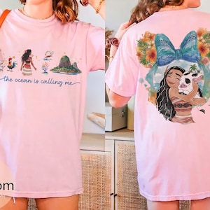 May include: Light pink t-shirt featuring a colorful graphic of a cartoon character and the text "the ocean is calling me". The back of the shirt has a graphic of the same character hugging a pig. The shirt is a casual style.