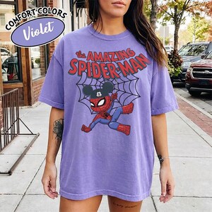 May include: A violet Comfort Colors t-shirt featuring a graphic of Spider-Man with a Mickey Mouse hat and the text "The Amazing Spider-Man" in red. The design includes a spider web. The shirt is a solid color.