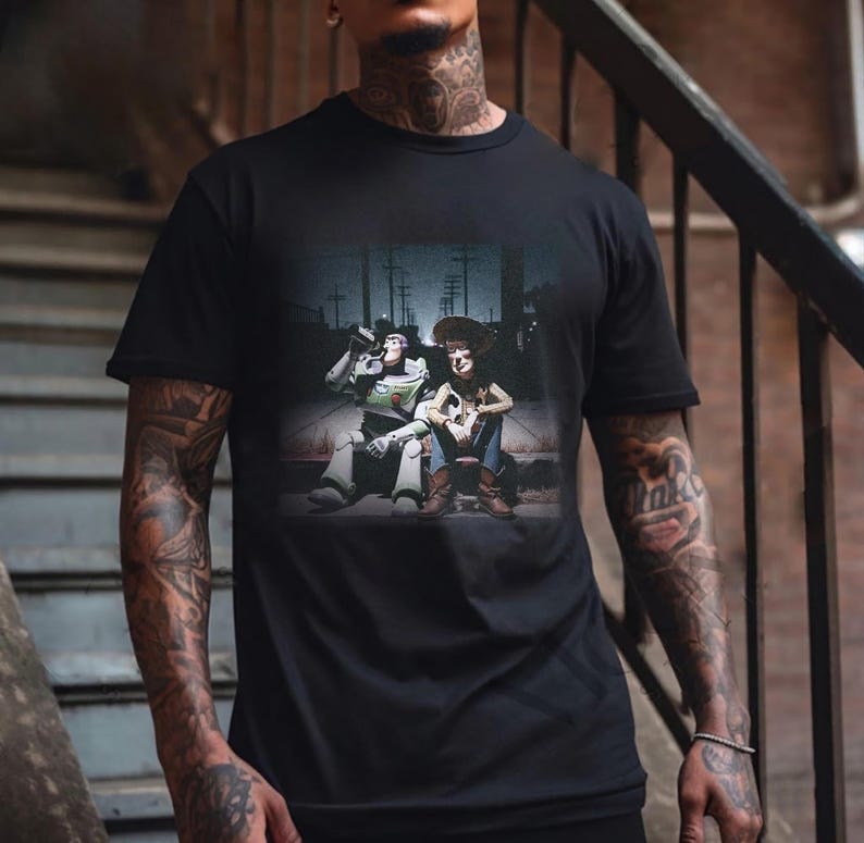 Puede incluir: Camiseta negra con un gr&aacute;fico de Buzz Lightyear y Woody de Toy Story. La imagen muestra a los personajes juntos, con Buzz bebiendo de una botella. El fondo representa una escena callejera.