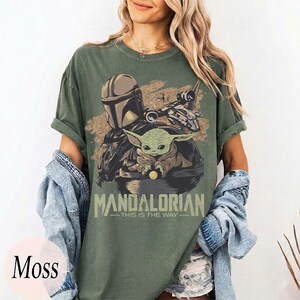 May include: Moss green t-shirt featuring a graphic of The Mandalorian and Grogu. The design includes the text "MANDALORIAN - THIS IS THE WAY" in a light green font. The shirt has short sleeves and a relaxed fit.