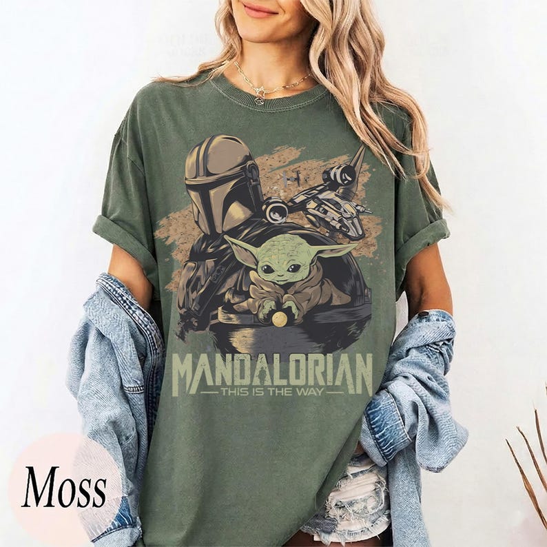 May include: Moss green t-shirt featuring a graphic of The Mandalorian and Grogu. The design includes the text "MANDALORIAN - THIS IS THE WAY" in a light green font. The shirt has short sleeves and a relaxed fit.