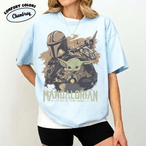 May include: Light blue t-shirt featuring a graphic of The Mandalorian and Grogu, with a spaceship. The text "MANDALORIAN - THIS IS THE WAY" is printed below the image. The shirt has short sleeves and a crew neck.