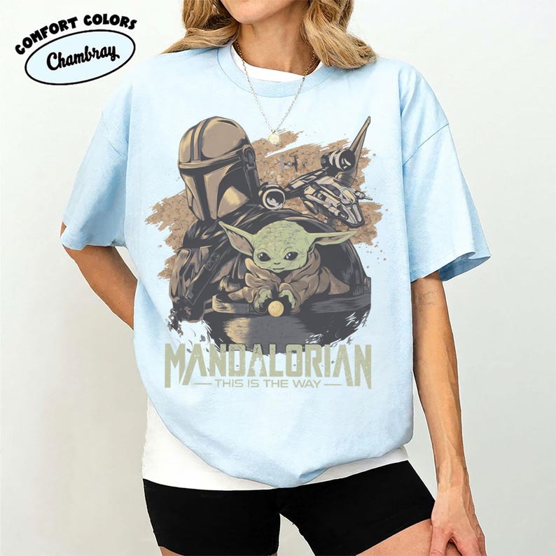 May include: Light blue t-shirt featuring a graphic of The Mandalorian and Grogu, with a spaceship. The text "MANDALORIAN - THIS IS THE WAY" is printed below the image. The shirt has short sleeves and a crew neck.