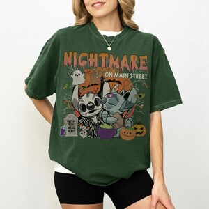 Puede incluir: Camiseta verde oscuro con un dise&ntilde;o de Halloween. El dise&ntilde;o presenta personajes de dibujos animados, calabazas y el texto "NIGHTMARE ON MAIN STREET". Una l&aacute;pida dice "WISH YOU WERE HERE!"