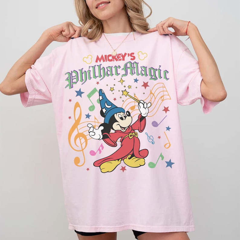 May include: A light pink t-shirt featuring Mickey Mouse as a sorcerer with a wand, musical notes, and stars. The text "Mickey's PhilharMagic" is printed above the graphic in colorful lettering. The shirt has short sleeves.
