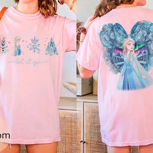 May include: Pink t-shirt featuring a watercolor design of characters and elements from a popular animated film. The front displays a snowman, princess, snowflake, and castle with the text "let it go." The back shows the princess in a blue dress.