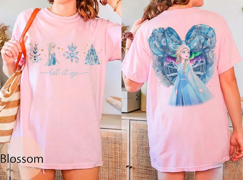 May include: Pink t-shirt featuring a watercolor design of characters and elements from a popular animated film. The front displays a snowman, princess, snowflake, and castle with the text "let it go." The back shows the princess in a blue dress.