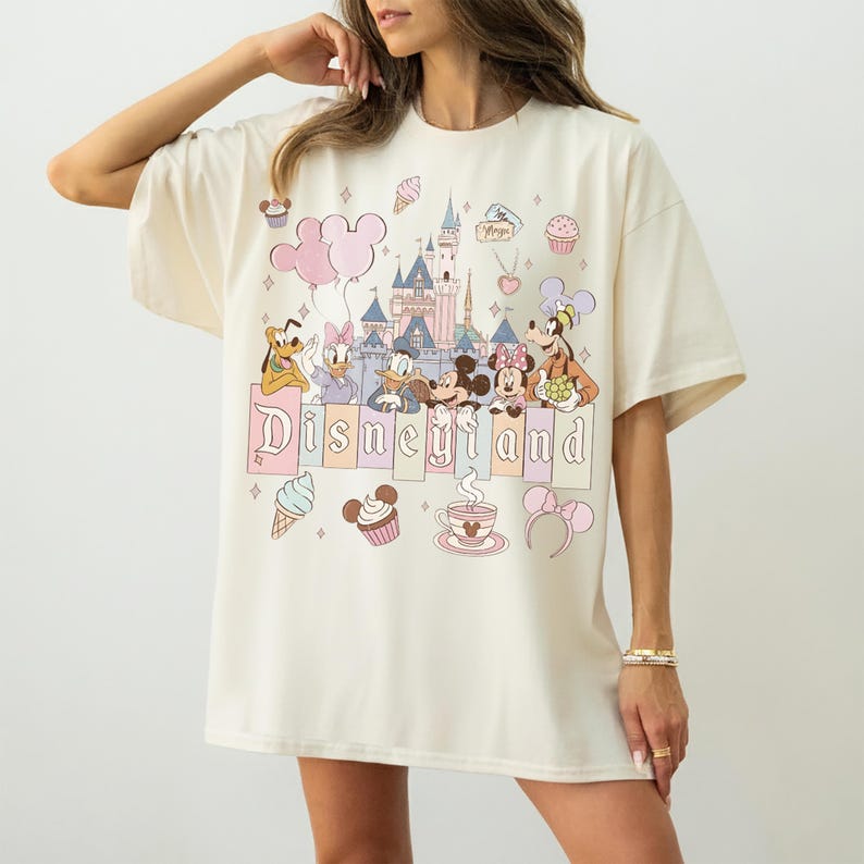 May include: Cream-coloured t-shirt with a colourful Disneyland graphic. The design includes Disney characters, a castle, balloons, and the word "Disneyland" in pastel colours. The shirt has short sleeves and a relaxed fit.