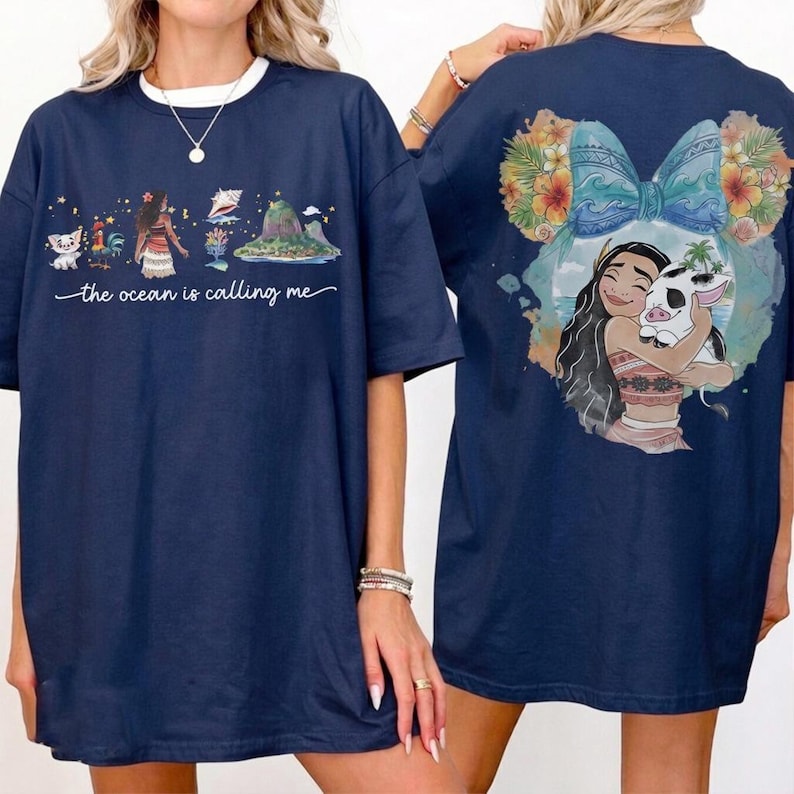 May include: Navy blue t-shirt featuring a front graphic with characters and the text "the ocean is calling me." The back of the shirt has a graphic of a character hugging a pig. The design includes floral and bow elements.